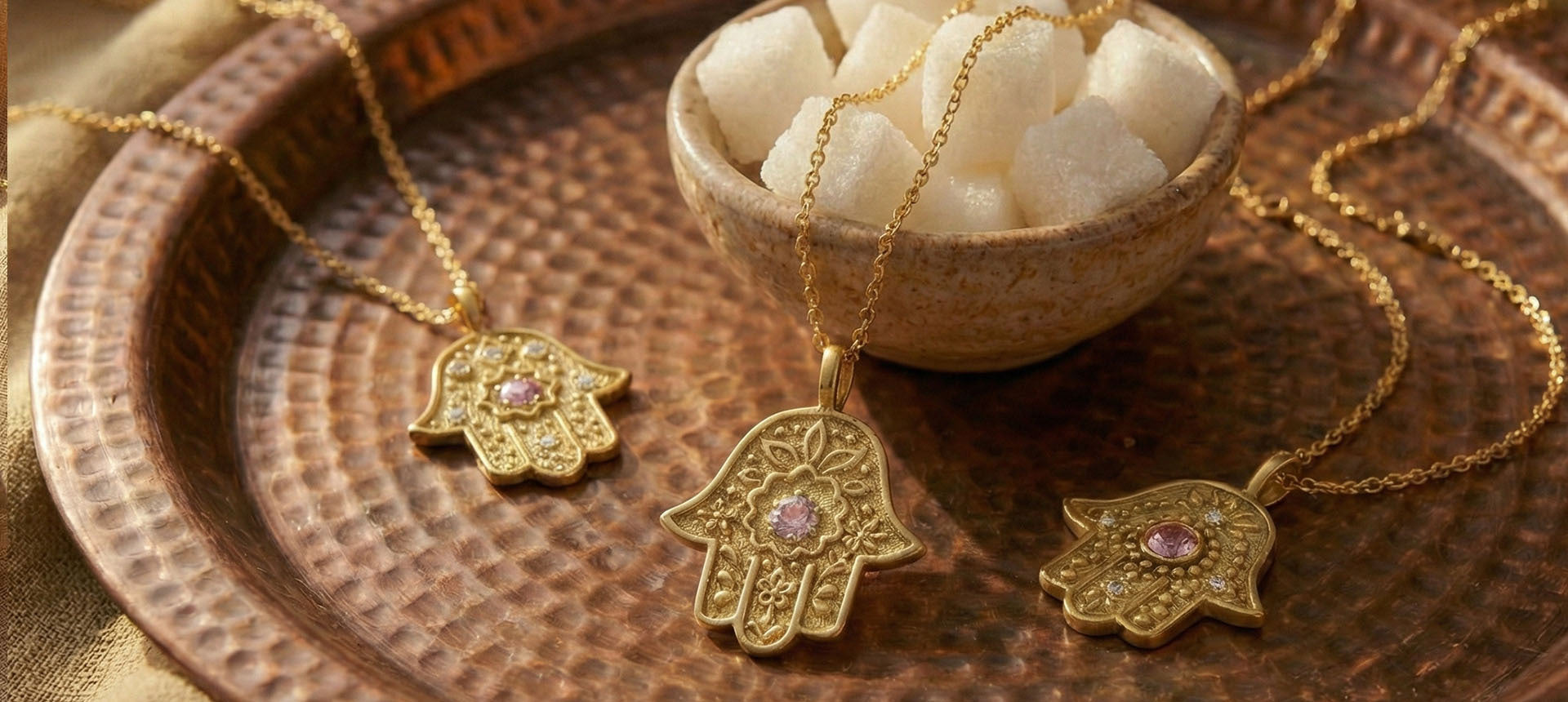 Four different gold Hamsa necklaces with pink sapphires arranged on a hammered copper tray with brown silk.