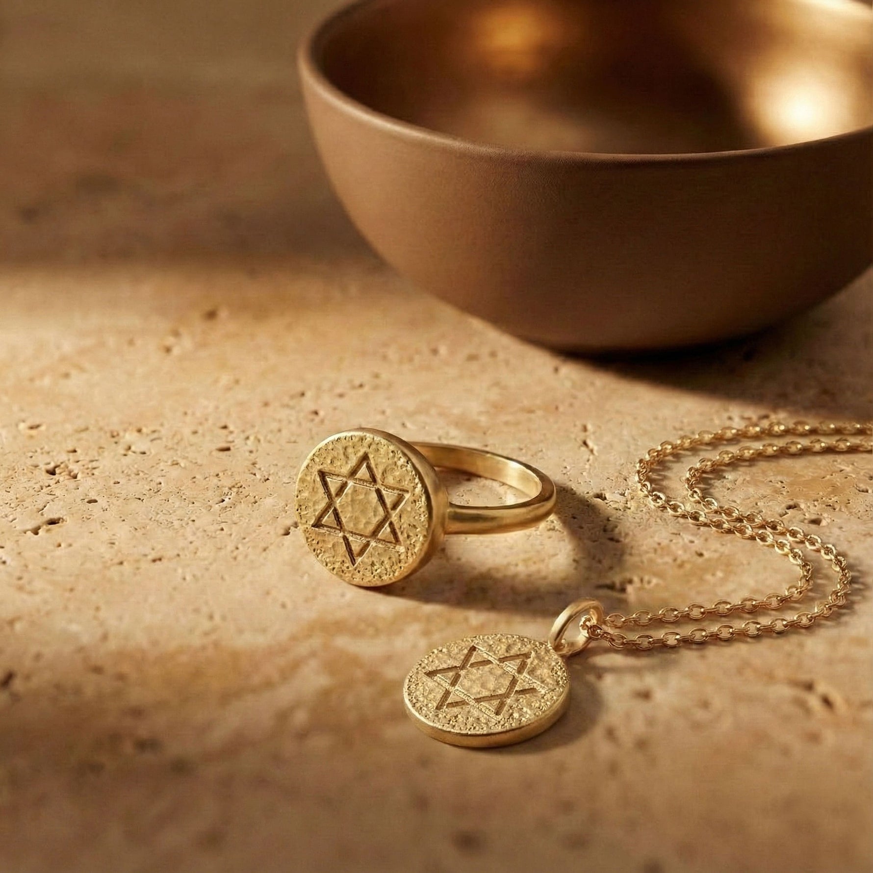 Classic Star of David coin  pendant necklace in gold displayed on a warm stone surface beside a bronze bowl.