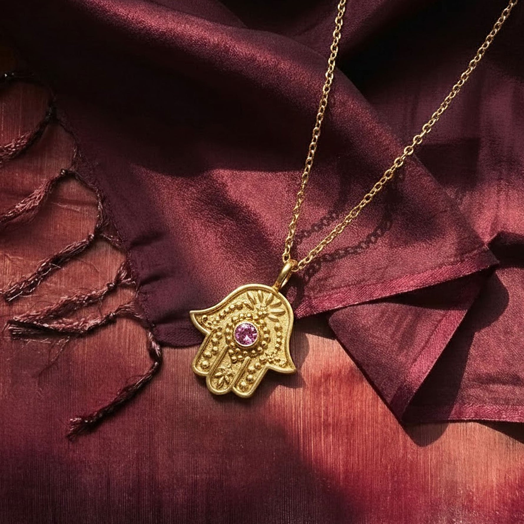 Gold Eternal Pulse Hamsa necklace with pink sapphire resting on draped burgundy silk fabric.