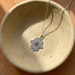 Silver necklace with a Hamsa hand pendant featuring a red gemstone, displayed on a textured beige surface.