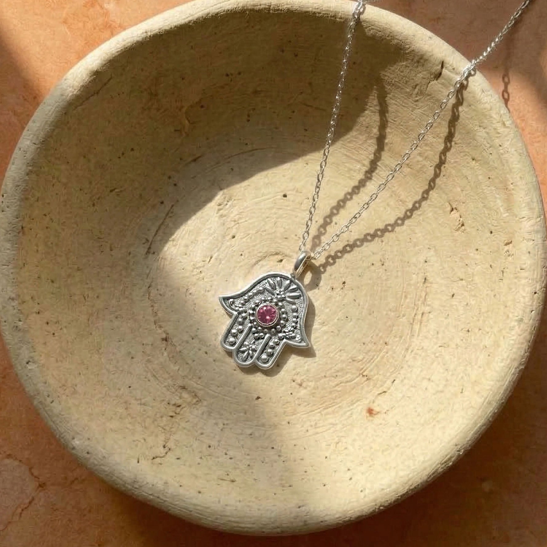 Silver necklace with a Hamsa hand pendant featuring a red gemstone, displayed on a textured beige surface.