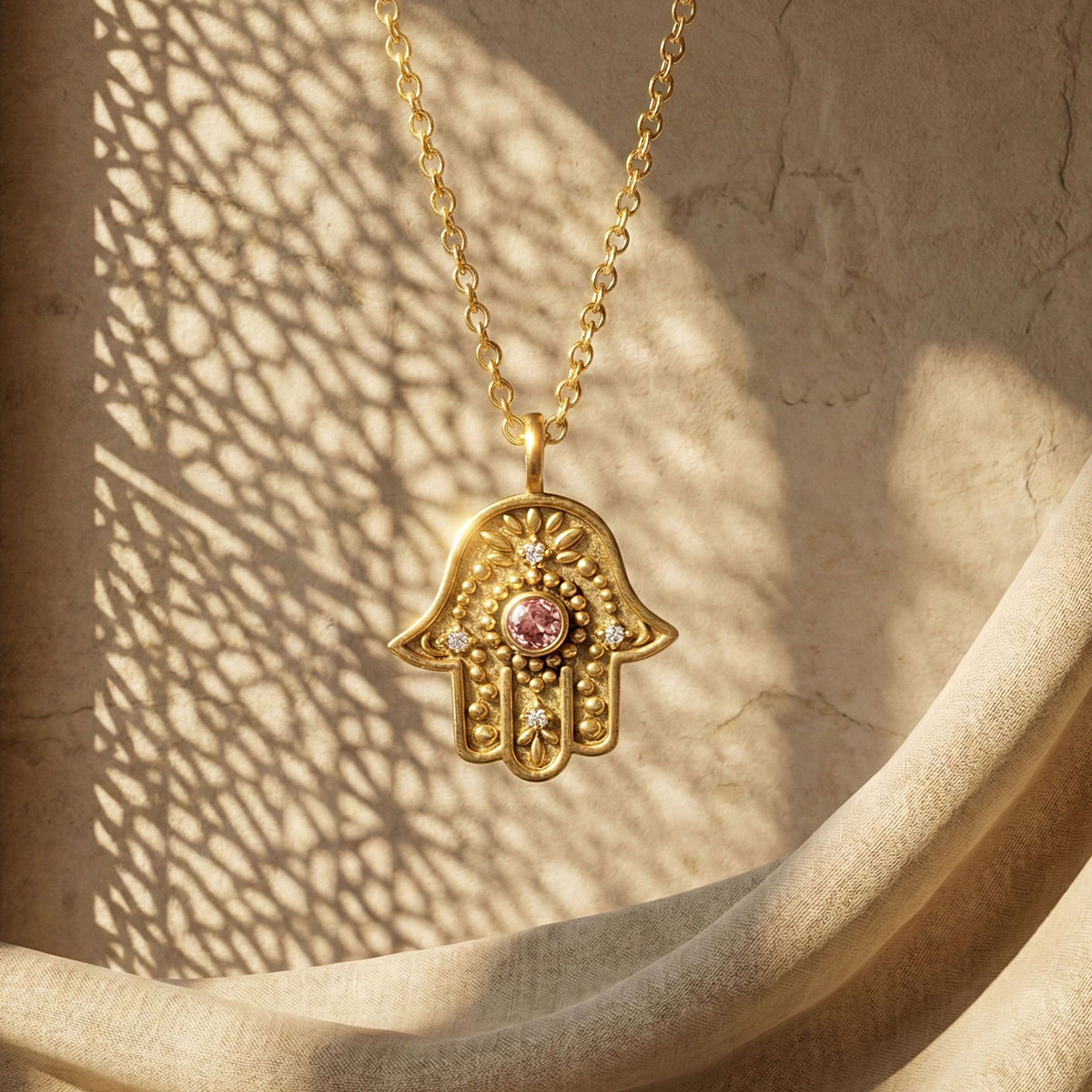 Gold Eternal Pulse Hamsa necklace with pink sapphire against a stone wall with lattice shadows.