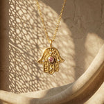 Gold Eternal Pulse Hamsa necklace with pink sapphire against a stone wall with lattice shadows.