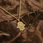 Gold Garden of Grace Hamsa necklace with pink sapphire resting on textured brown silk.