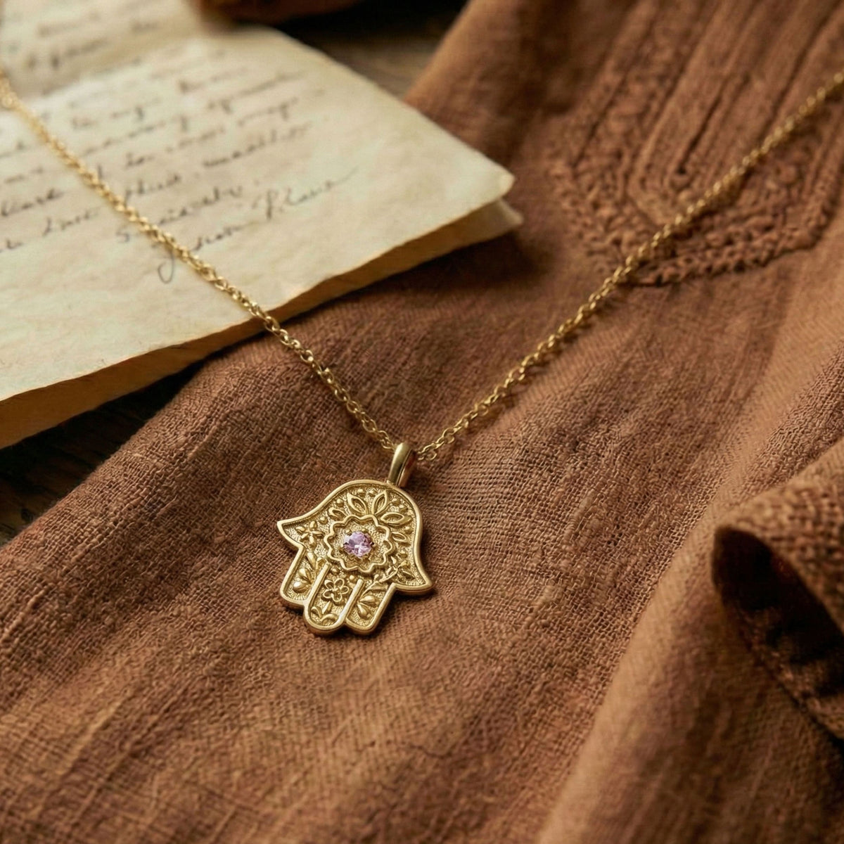 Gold Hamsa necklace with floral motif resting on a brown linen shirt next to an old handwritten letter.