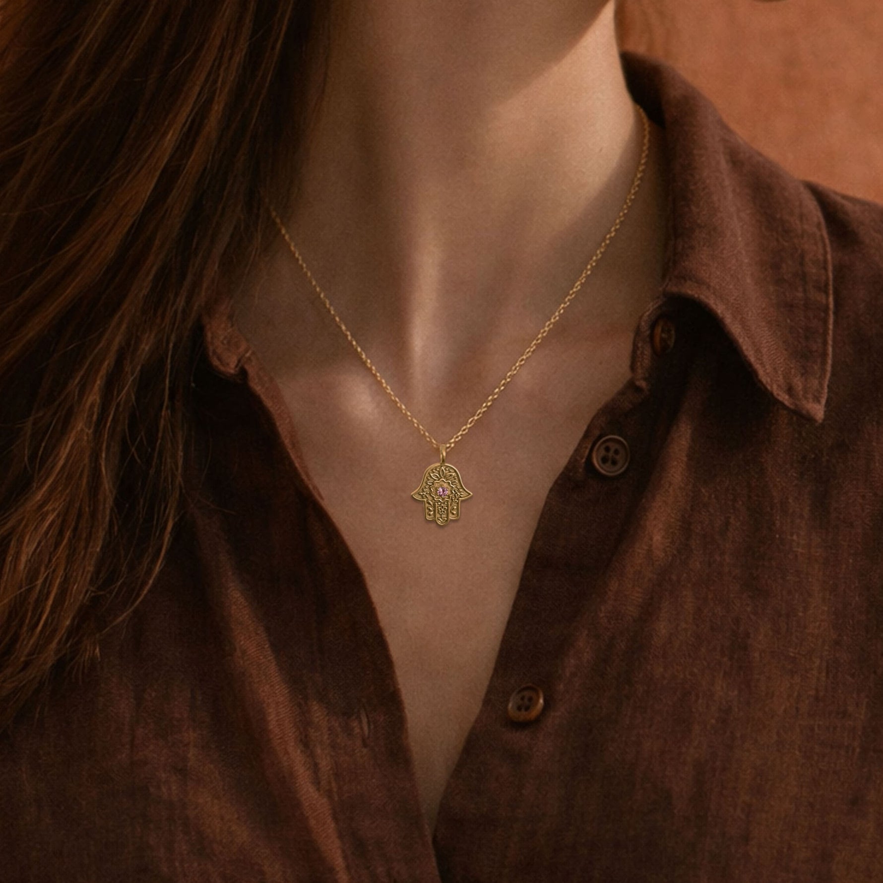Woman wearing the gold Garden of Grace Hamsa necklace over a dark brown linen shirt.