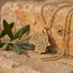 Gold Garden of Grace Hamsa necklace with pink sapphire resting on stone next to an olive branch.