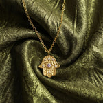 Gold Radiant Anchor Hamsa necklace resting on draped olive green silk fabric.