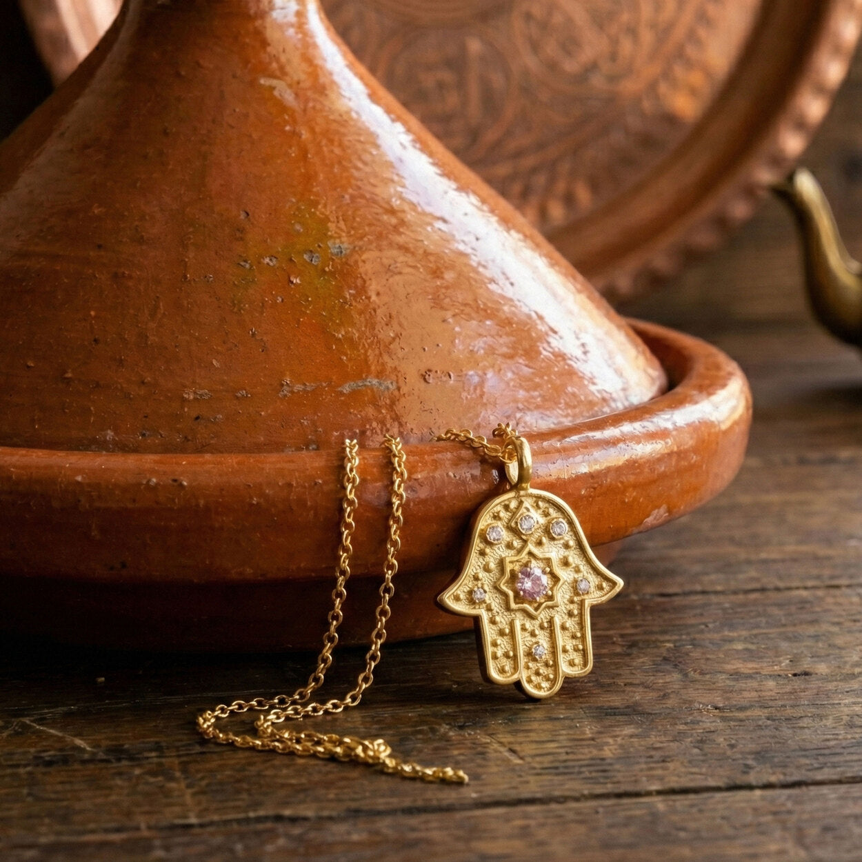 Gold Hamsa necklace with pink sapphire and diamonds leaning against a clay tajine.
