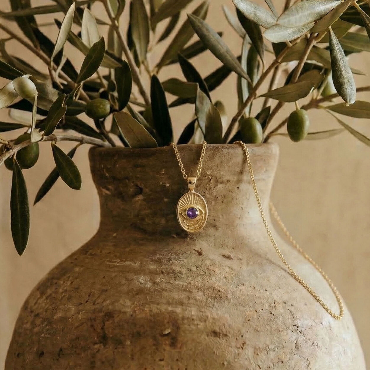 The Window to the Soul necklace draped over an ancient-style ceramic vase with olive branches, representing the brand's Tel Aviv atelier roots.